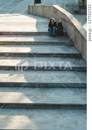 Woman with suitcase sitting on wide concrete stairs waiting Woman with suitcase sitting on wide concrete stairs waiting 127502722