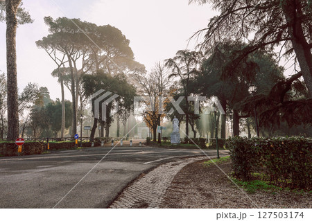Serene scene of tranquil park surrounded by tall trees and prominent statue. Soft morning light filters through leaves, creating peaceful atmosphere. Villa Borghese gardens, Rome, Italy Serene scene of tranquil park surrounded by tall trees and prominent statue. Soft morning light filters through leaves, creating peaceful atmosphere. Villa Borghese gardens, Rome, Italy 127503174