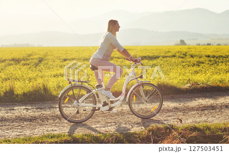 Young woman rides electric bicycle on dusty country road, view from side, sun backlight field with yellow flowers background 127503451