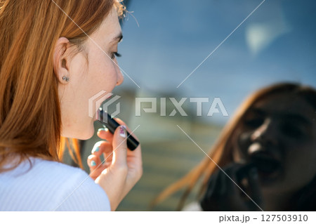 Closeup of a young redhead woman driver correcting her makeup with dark red lipstick looking in car reflective window. 127503910