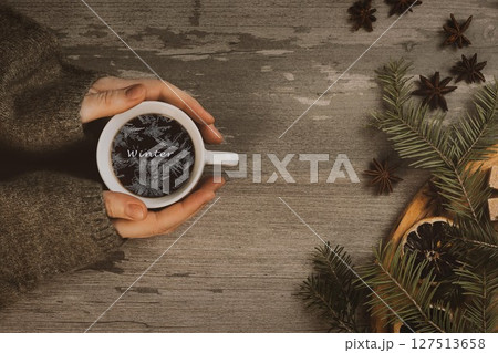 Christmas atmospheric set. Female hands in a cozy green sweaters sleeves with coffee cup and frosty lettering in it. Spruce twigs, anise on a wooden table. Christmas atmospheric set. Female hands in a cozy green sweaters sleeves with coffee cup and frosty lettering in it. Spruce twigs, anise on a wooden table. 127513658