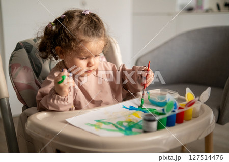Side view of a young girl using a paintbrush with gouache while seated at a small art table. Childhood creativity in action. Side view of a young girl using a paintbrush with gouache while seated at a small art table. Childhood creativity in action. 127514476
