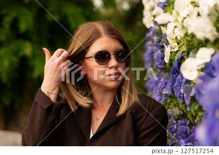 Young stylish woman poses outdoors near vibrant floral backdrop. Young stylish woman poses outdoors near vibrant floral backdrop. 127517254