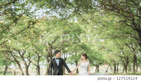 公園の並木道を手を繋いで歩くドレス姿のカップル（ウェディング・ウェディングフォト・新郎新婦） 127519124