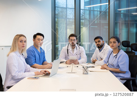 A diverse group of medical professionals sitting at a table in a modern office, engaged in a meeting. A diverse group of medical professionals sitting at a table in a modern office, engaged in a meeting. 127520887