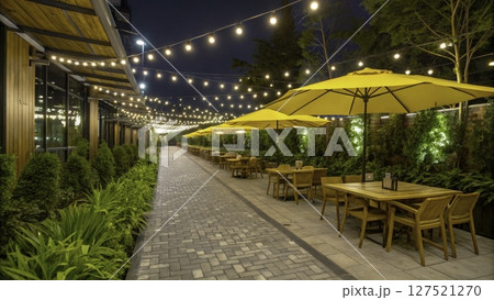 Outdoor dining area adorned with yellow umbrellas and decorative lights creates an inviting evening atmosphere in a landscaped setting 127521270