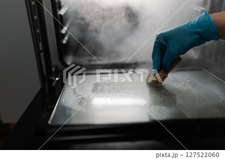 Cropped shot of cleaning company worker male wearing blue glove using sponge for cleaning glass door of gas oven in dark kitchen room. Concept of hygiene and cleanliness in domestic environment. Cropped shot of cleaning company worker male wearing blue glove using sponge for cleaning glass door of gas oven in dark kitchen room. Concept of hygiene and cleanliness in domestic environment. 127522060