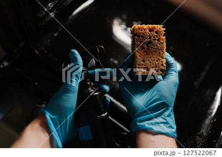 High-angle view of housewife in gloves spraying detergent on sponge, preparing to clean glass door of modern oven in kitchen with dark interior. Concept of cleanliness during routine chores. High-angle view of housewife in gloves spraying detergent on sponge, preparing to clean glass door of modern oven in kitchen with dark interior. Concept of cleanliness during routine chores. 127522067