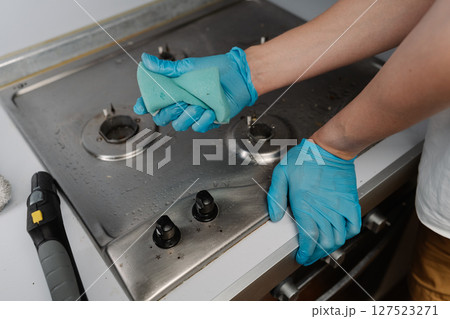 Cleaning service employee wearing blue gloves using steam cleaner and sponge to remove grease and dirt from stainless steel gas stove, ensuring hygiene and cleanliness in kitchen, close-up. 127523271