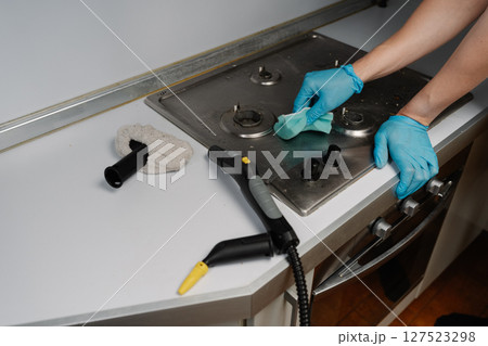 Cropped shot of housekeeper wearing blue gloves using steam cleaner and sponge to remove grease and dirt from stainless steel gas stove, ensuring hygiene and cleanliness in kitchen, close-up. Cropped shot of housekeeper wearing blue gloves using steam cleaner and sponge to remove grease and dirt from stainless steel gas stove, ensuring hygiene and cleanliness in kitchen, close-up. 127523298
