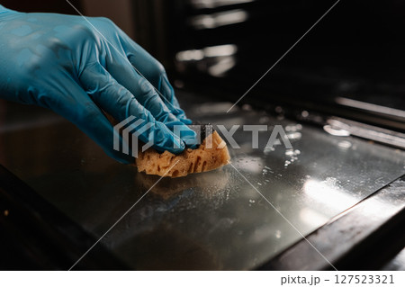 Detail cropped shot of unrecognizable man wearing blue gloves wiping glass door of oven with sponge, ensuring thorough cleaning and maintenance of kitchen appliances. Concept of hygiene, cleanliness. 127523321