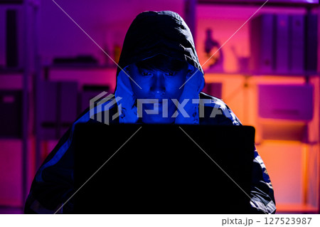 Young Man in Hoodie Working on Laptop in Dark Tech Environment 127523987
