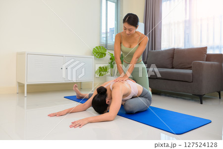 Two sporty girls practice yoga with partner at home. Trainer helps student to do seated forward fold exercise Two sporty girls practice yoga with partner at home. Trainer helps student to do seated forward fold exercise 127524128