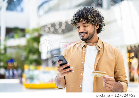 A smiling man in a shopping mall, holding a phone and a credit card, ready to make a purchase, depicting the convenience of online shopping. 127524458