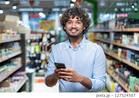 A smiling man in a supermarket, holding a phone, looking at the camera. Shelves with products are in the background. A smiling man in a supermarket, holding a phone, looking at the camera. Shelves with products are in the background. 127524507