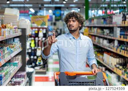 A man shops in a grocery store, extending his finger as he looks toward the shelves, pushing a shopping cart filled with items. A man shops in a grocery store, extending his finger as he looks toward the shelves, pushing a shopping cart filled with items. 127524509