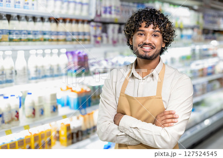 A cheerful store employee stands with folded arms, smiling in front of the dairy section filled with products. The grocery store worker looks confident and ready to assist. A cheerful store employee stands with folded arms, smiling in front of the dairy section filled with products. The grocery store worker looks confident and ready to assist. 127524545