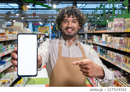 A smiling supermarket employee holds up a phone with a blank screen, inviting customers to scan, or use an app. 127524562