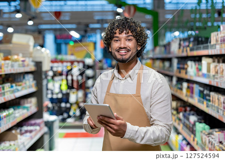 A smiling grocery store worker holds a tablet while standing in an aisle, surrounded by products on shelves. 127524594