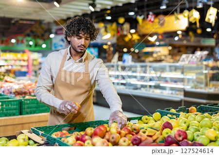 A grocery worker wearing an apron and gloves carefully selects apples from a fresh produce display, checking for quality. 127524614