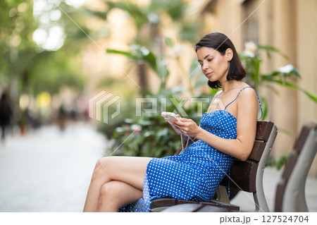 Young woman with phone sitting on bench in street Young woman with phone sitting on bench in street 127524704