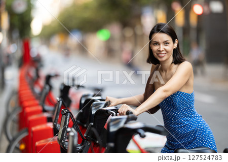Brunette in blue dress rents electric bike on street of european city 127524783