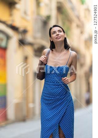 Cheerful brunette girl in blue dress walking along the street among architecture with bag 127524901
