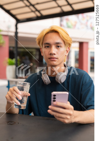 Asian gay teenager drinking water and using phone outdoors in cafe restaurant Asian gay teenager drinking water and using phone outdoors in cafe restaurant 127525669