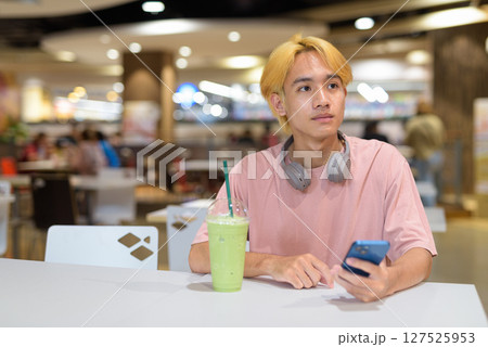 Thai gay teen man with headphones using phone and drinking iced latte green tea in modern cafe 127525953