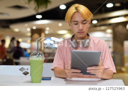 Thai gay teen man with headphones using digital tablet and drinking iced latte green tea 127525963