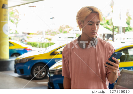 Young Asian gay teen tourist using phone outdoors with taxis behind Young Asian gay teen tourist using phone outdoors with taxis behind 127526019