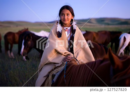 Girl in Traditional Attire Riding a Horse Among a Group of Horses at Sunset in an Open Field 127527820