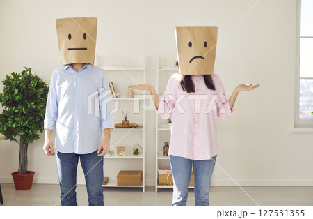 Faceless man and woman with paper bags on head with confused faces standing in living room at home. 127531355