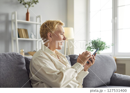 Cheerful mature woman sitting on sofa, scrolling social media and enjoying online entertainment. 127531400