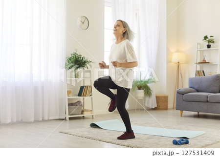 Smiling mature woman running in place in living room, following video tutorial for home workout. Smiling mature woman running in place in living room, following video tutorial for home workout. 127531409