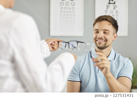 Optician, optometrist or ophthalmologist giving new glasses to young smiling man with vision problem Optician, optometrist or ophthalmologist giving new glasses to young smiling man with vision problem 127531431