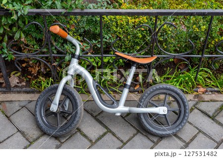 A compact metal balance bike for children parked against a wrought iron fence in a neatly landscaped area with paving and greenery. A compact metal balance bike for children parked against a wrought iron fence in a neatly landscaped area with paving and greenery. 127531482