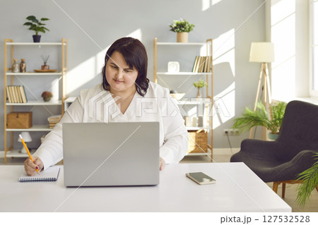 Woman studying online, sitting at desk at home, using laptop and writing in notebook Woman studying online, sitting at desk at home, using laptop and writing in notebook 127532528