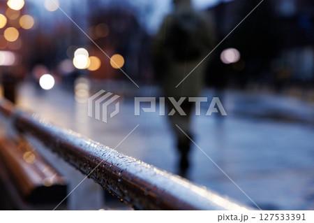 Raindrops glisten on a railing as a person walks through a vibrant, bustling urban street at dusk Raindrops glisten on a railing as a person walks through a vibrant, bustling urban street at dusk 127533391