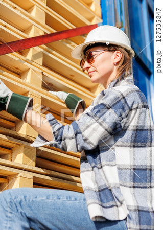Portrait happy female driver wearing sunglasses and safety helmet smiles in front of blue truck and stacked pallets. Female person girl professional job career working driving freight engine lorry 127535847