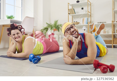 Funny couple in bright sportswear doing sport exercises at home lying on yoga mat with dumbbells. Funny couple in bright sportswear doing sport exercises at home lying on yoga mat with dumbbells. 127536562