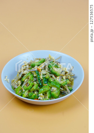 A vibrant dish of stir-fried bitter melon with anchovies and eggs, served in a white bowl on a light orange background. A vibrant dish of stir-fried bitter melon with anchovies and eggs, served in a white bowl on a light orange background. 127536575
