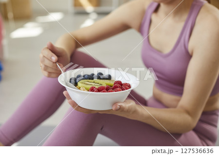 Fit young woman in sportswear holding bowl and eating cereal with fruit and berries Fit young woman in sportswear holding bowl and eating cereal with fruit and berries 127536636