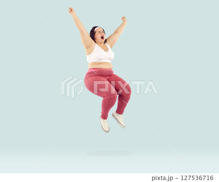 Fat excited happy woman wearing sportswear jumping isolated on studio blue background. 127536716