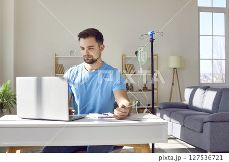 Young man working on a laptop at the desk at home or in office while receiving IV vitamin therapy. 127536721