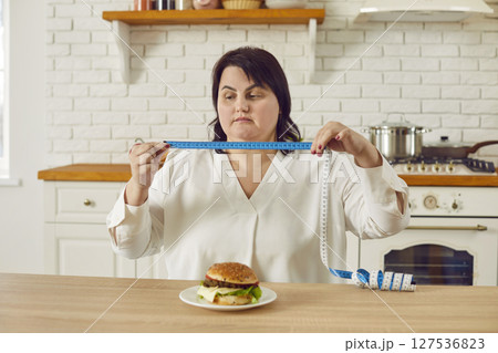 Fat overweight young woman sitting on kitchen holding a measuring tape with a burger on the table. 127536823