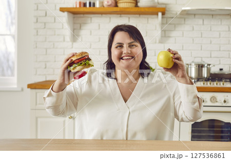 Young overweight plus size fat woman holding a burger in one hand and an apple in other. Young overweight plus size fat woman holding a burger in one hand and an apple in other. 127536861