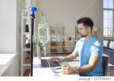 Young man working on a laptop at the desk at home or in office while receiving IV vitamin therapy. 127536867