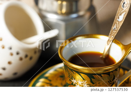 Elegant coffee cup with golden details sits on a vibrant saucer beside a stylish coffee maker in cozy setting Elegant coffee cup with golden details sits on a vibrant saucer beside a stylish coffee maker in cozy setting 127537530