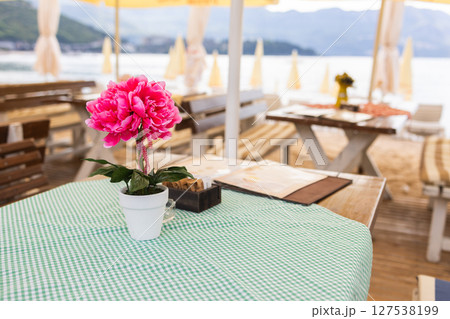 Seaside cafe table with flower decoration and beach view. Concept of coastal relaxation, vacation ambiance and summer leisure by the sea. Seaside cafe table with flower decoration and beach view. Concept of coastal relaxation, vacation ambiance and summer leisure by the sea. 127538199